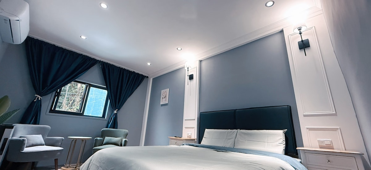 A serene bedroom space is feature with a spacious bed and two accent chairs. Soft blue walls are complemented by dark curtains and soft lighting from recessed ceiling fixtures. A window allows natural light, and a small nightstand sits beside the bed.
