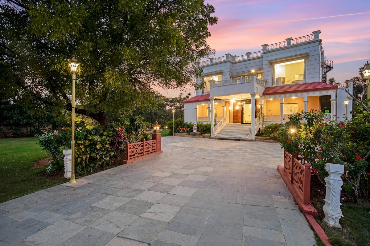A spacious villa is depicted at dusk, featuring a grand facade with multiple steps leading to the entrance. Surrounding gardens are lush and well-maintained, with outdoor lighting enhancing the serene atmosphere. The evening sky casts a soft glow over the charming architecture.