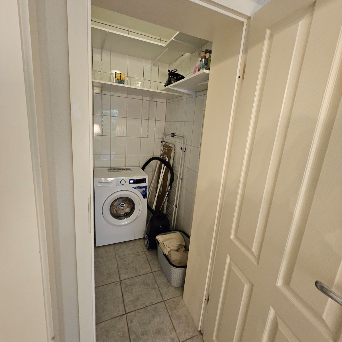 A laundry space is fitted with a washing machine and various storage items. Shelving is utilized for organizing cleaning supplies, while a laundry basket and a vacuum cleaner are also present. The tiled floor complements the clean and functional layout of the area.