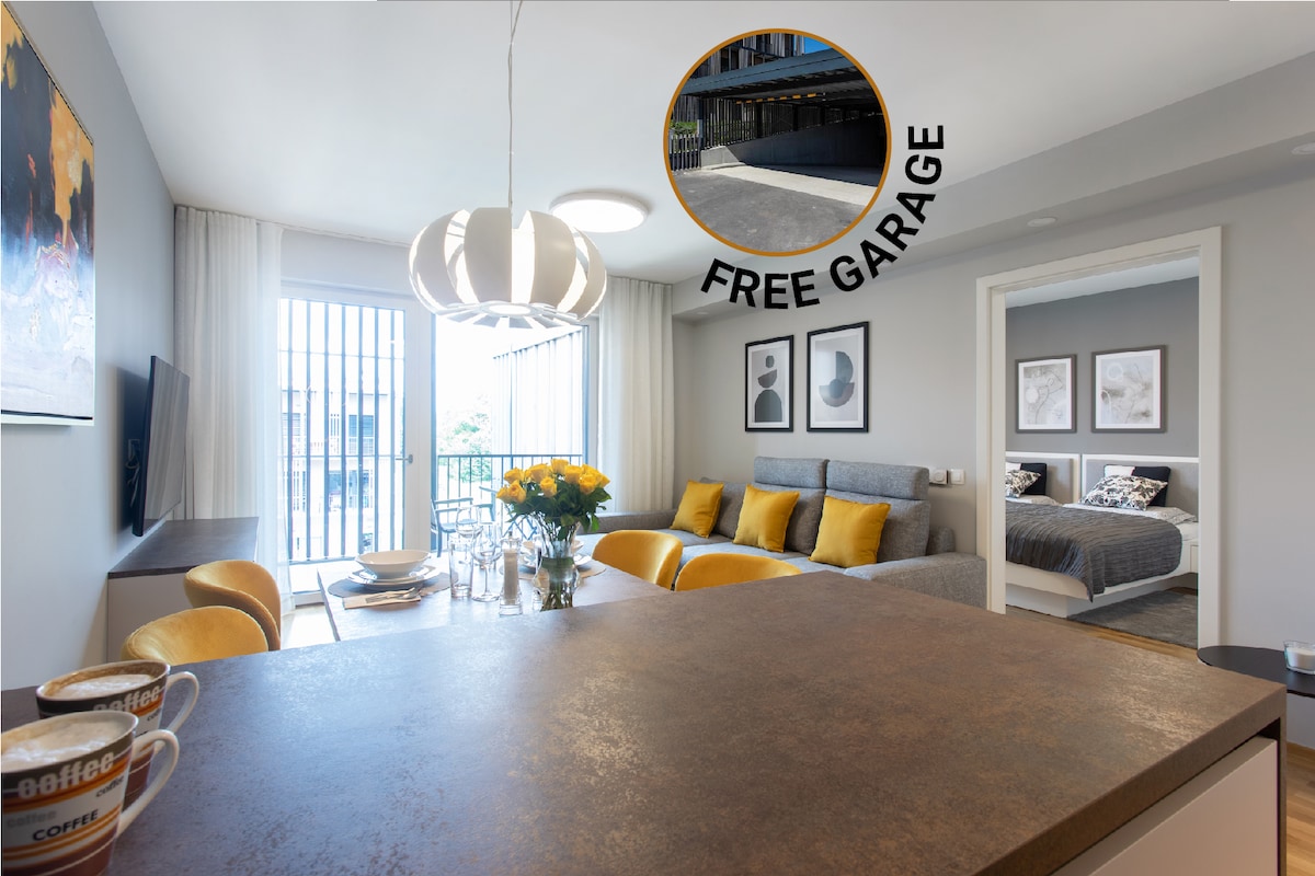 The spacious living area features a modern design with a comfortable gray sofa and a dining table set for four. Natural light fills the space through large glass doors leading to a balcony. Bright yellow accents add a cheerful touch, complemented by a kitchen island visible in the foreground.