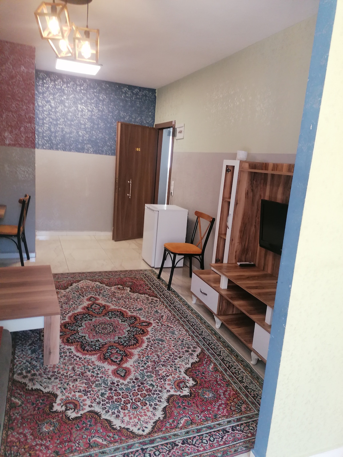 A small living area is depicted, featuring a patterned rug laid over tiled flooring. A wooden coffee table sits in front of a low TV unit with a television. A white refrigerator is positioned beside the door, and two chairs are placed at a nearby table.