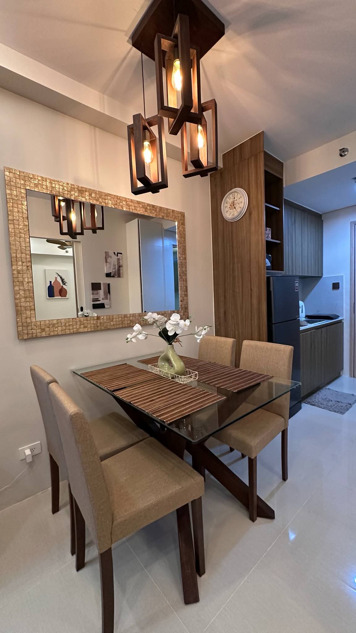 A dining area features a glass table surrounded by four beige chairs. A decorative mirror is mounted on the wall, reflecting warm light from two hanging fixtures. A small vase with white flowers is placed at the table's center. The kitchen area is visible in the background.