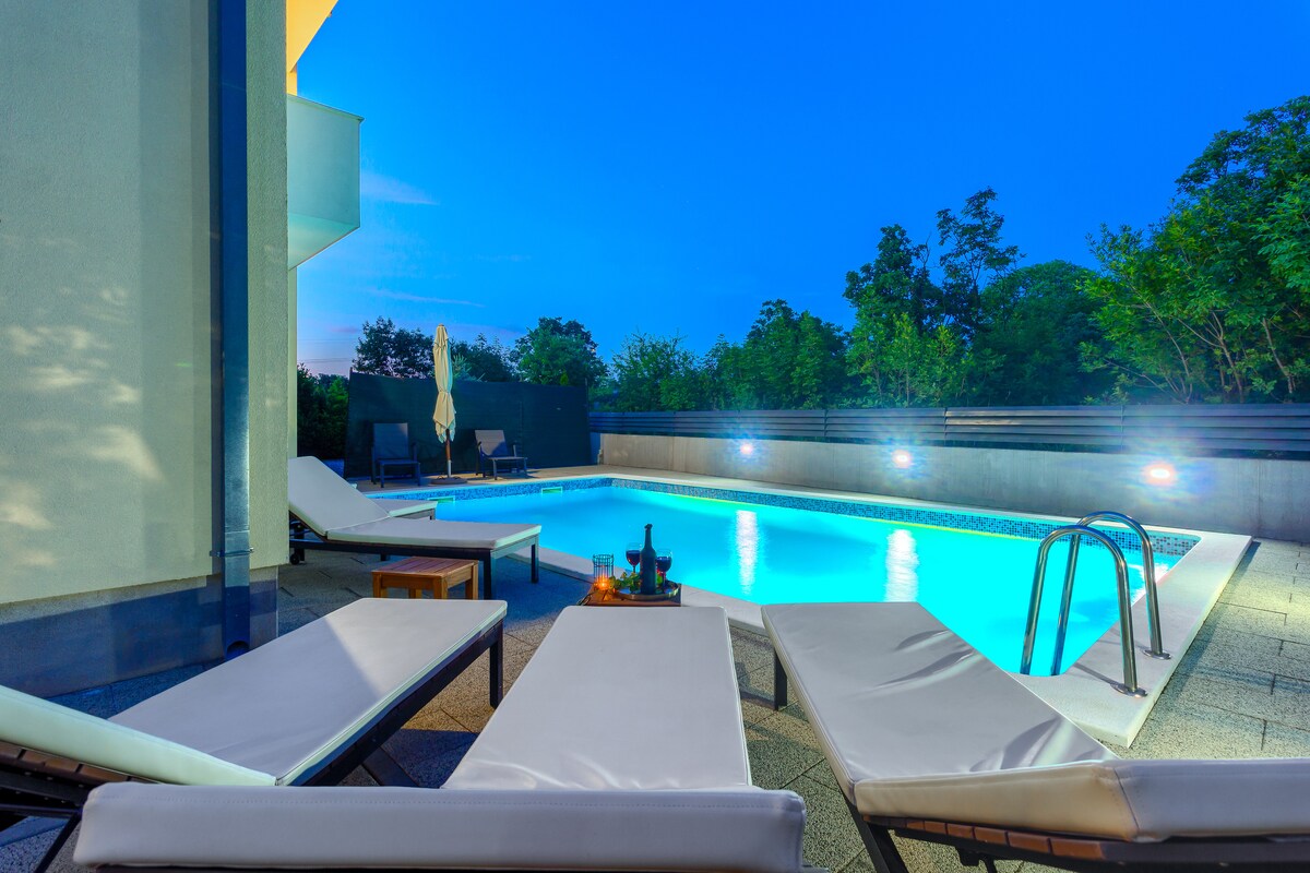 An inviting outdoor pool area is surrounded by lush greenery, with several loungers arranged beside the water. A small table sits near the edge, complementing the tranquil atmosphere created by the soft evening light reflecting off the pool's surface.