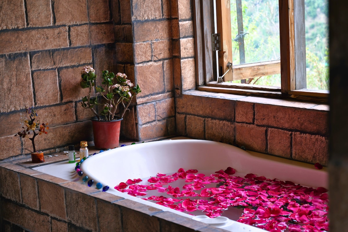 A sunlit bathroom features a freestanding bathtub filled with pink rose petals. Natural light filters through a wooden window, highlighting the textured stone walls. A small potted plant and decorative items are placed on the edge of the tub, creating a serene setting.