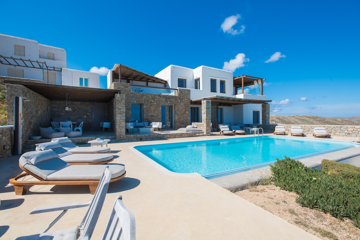 A spacious outdoor area features a large infinity pool surrounded by sun loungers and lounge chairs. Various outdoor seating areas provide great views of the landscape, complemented by a modern villa structure with multiple levels and ample windows.