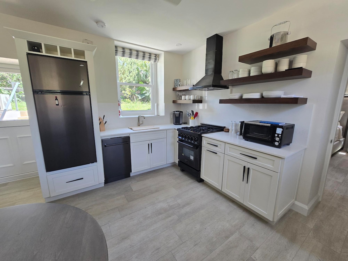 A modern kitchen is equipped with a black refrigerator, a dishwasher, and an oven range. White cabinetry contrasts with dark wooden shelves, which are arranged with plates and glassware. A window allows natural light to brighten the space, enhancing functionality.