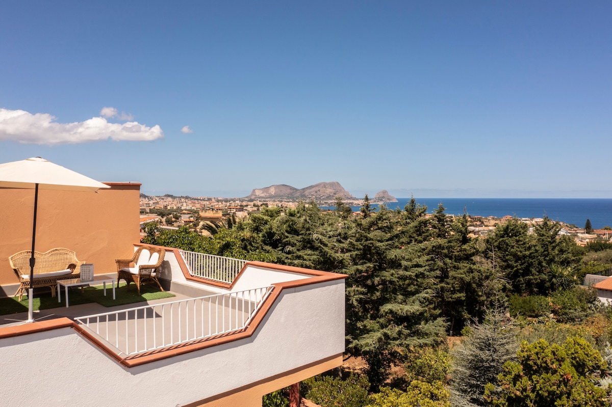 A scenic terrace offers a panoramic view of the lush landscape and coastline. A shaded seating area, complete with wicker chairs and an umbrella, invites relaxation while the blue sea and distant mountains create a tranquil backdrop.