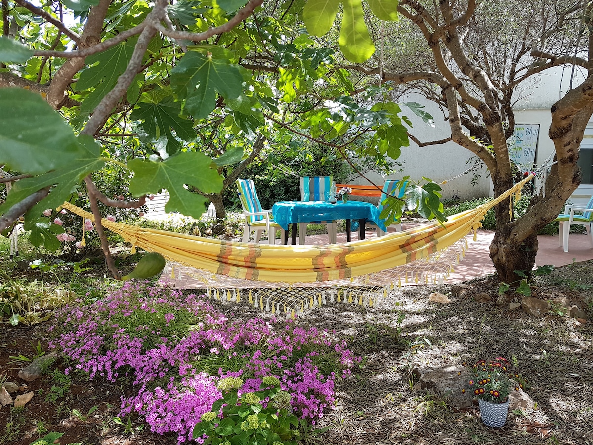 A vibrant garden area is depicted, featuring a yellow hammock gently swaying between two trees. A round table set with blue and green chairs is visible nearby, surrounded by blooming pink flowers and lush greenery.