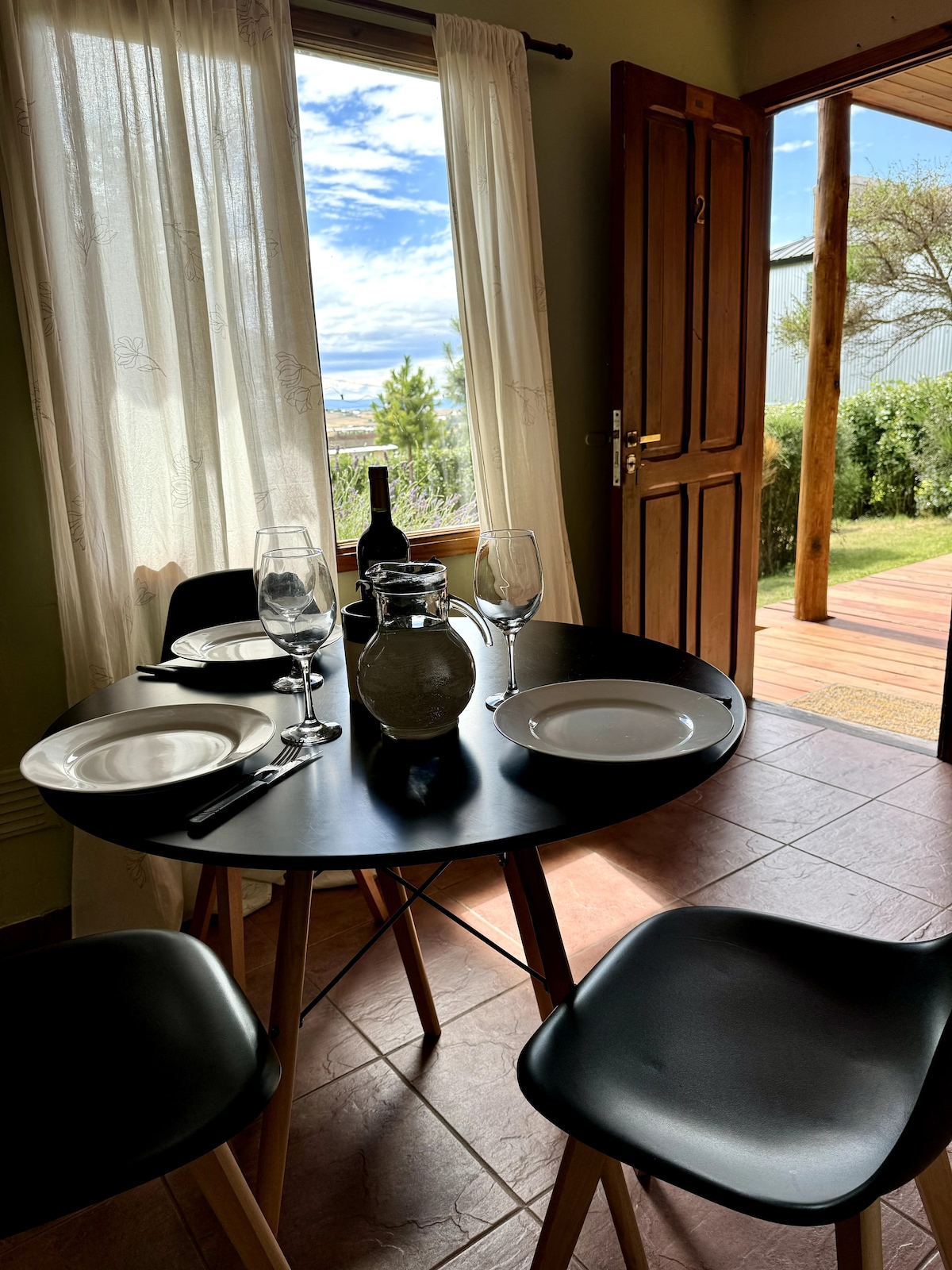 A dining area is showcased, featuring a round black table set with plates, glasses, and cutlery. Natural light enters through a window dressed with sheer curtains, revealing a glimpse of the outdoor greenery. A wooden door stands open, enhancing the sense of flow between spaces.