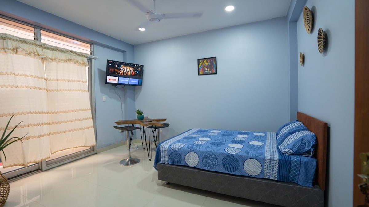 A private room features a spacious bed covered with a blue patterned blanket. A mounted smart TV displays information on a wall. Two small tables with chairs are positioned beside a window, partially covered by light curtains. A ceiling fan is present to circulate air.