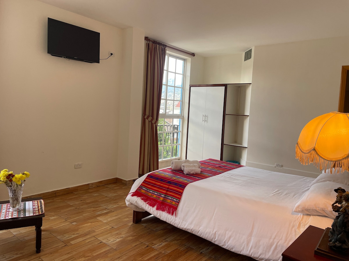 A well-lit private room is furnished with a spacious bed, adorned with a colorful woven blanket. A flat-screen television is mounted on the wall. Natural light enters through a large window, with views of the surrounding area. A decorative lamp and a small table with flowers enhance the ambiance.