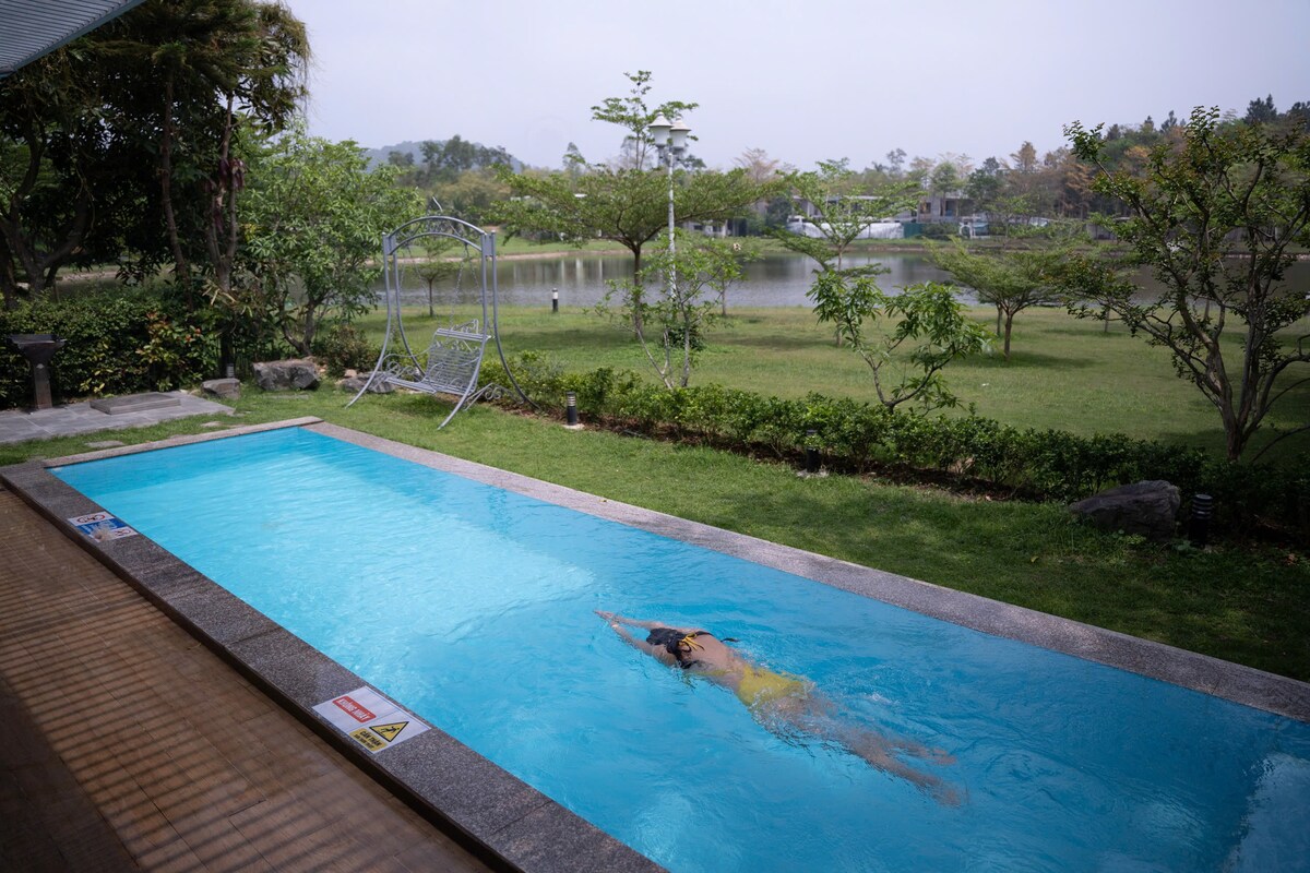 A serene swimming pool is set within a lush garden, featuring clear blue water. A swimmer glides through the pool, while vibrant greenery and trees surround the area. A peaceful lake can be seen in the background, contributing to the tranquil atmosphere.