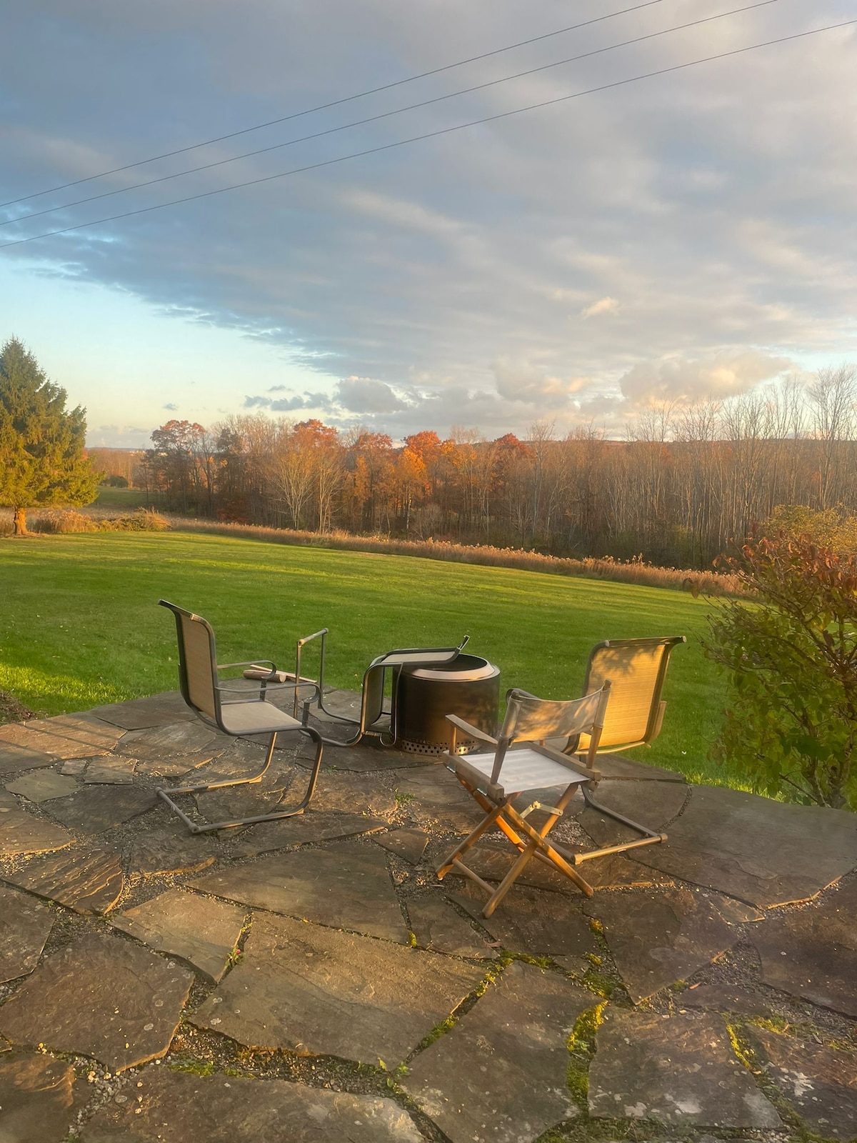 A stone patio offers a view of the expansive green lawn and tree line, showcasing fall foliage. Two chairs are positioned around a firepit, inviting relaxation while enjoying the scenic landscape and changing colors of the trees against a softly illuminated sky.