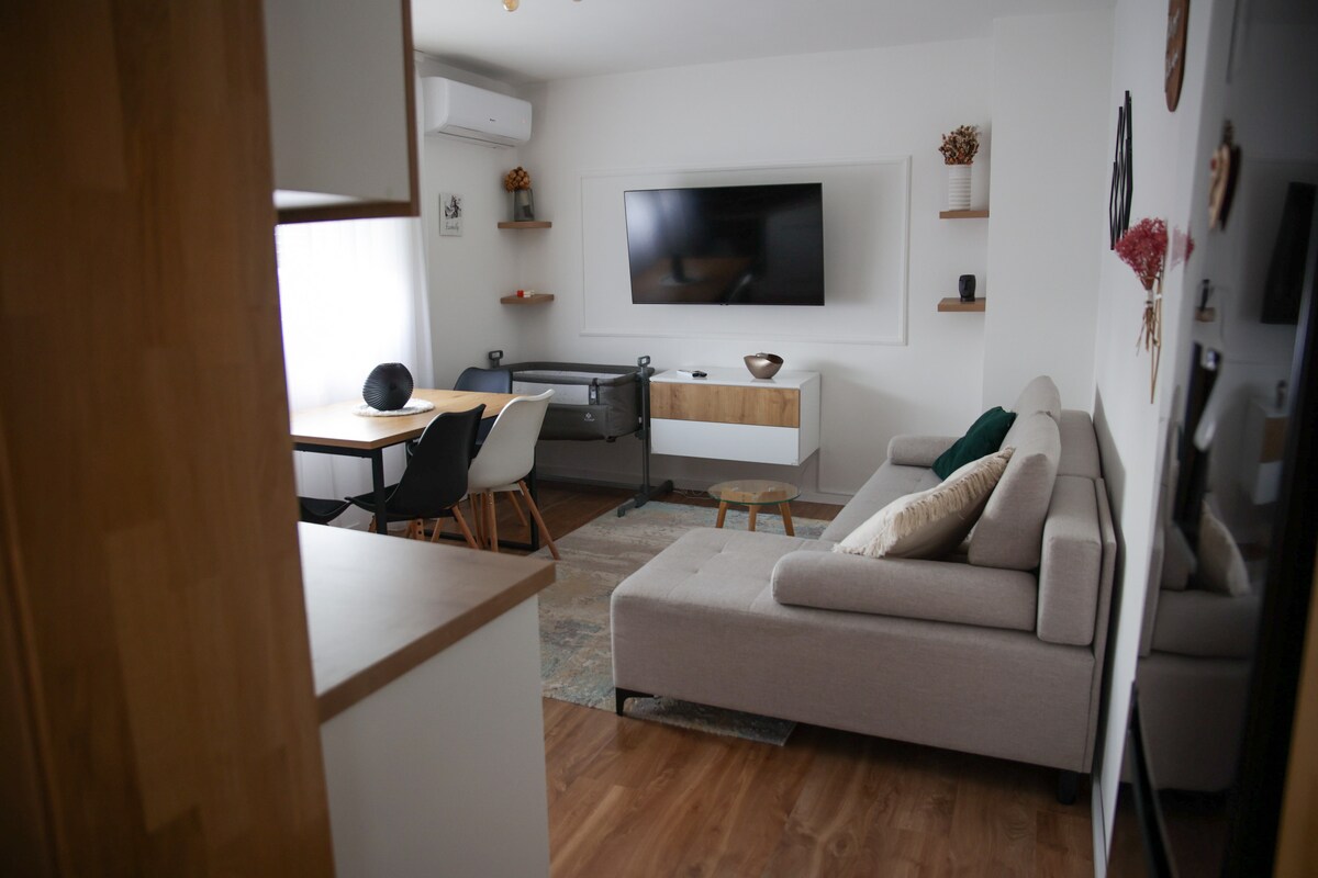 A modern living area features a spacious sectional sofa and a wooden dining table with a few chairs. A television mounted on the wall is surrounded by decorative shelving, while natural light filters through a sheer curtain, creating a bright and welcoming environment.