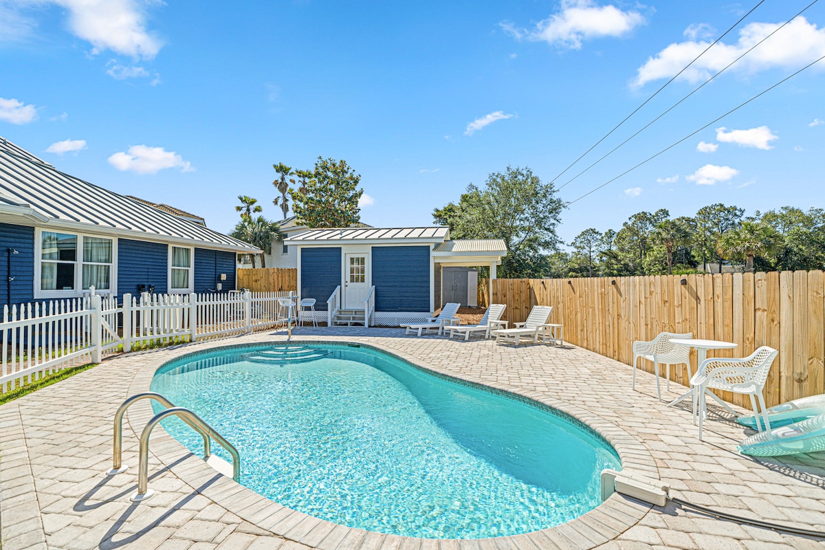 A private pool is surrounded by a spacious paved area, featuring lounge chairs for relaxation. A wooden fence provides privacy, while the vibrant blue exterior of the house contrasts with the greenery in the background. Clear skies highlight the inviting atmosphere.