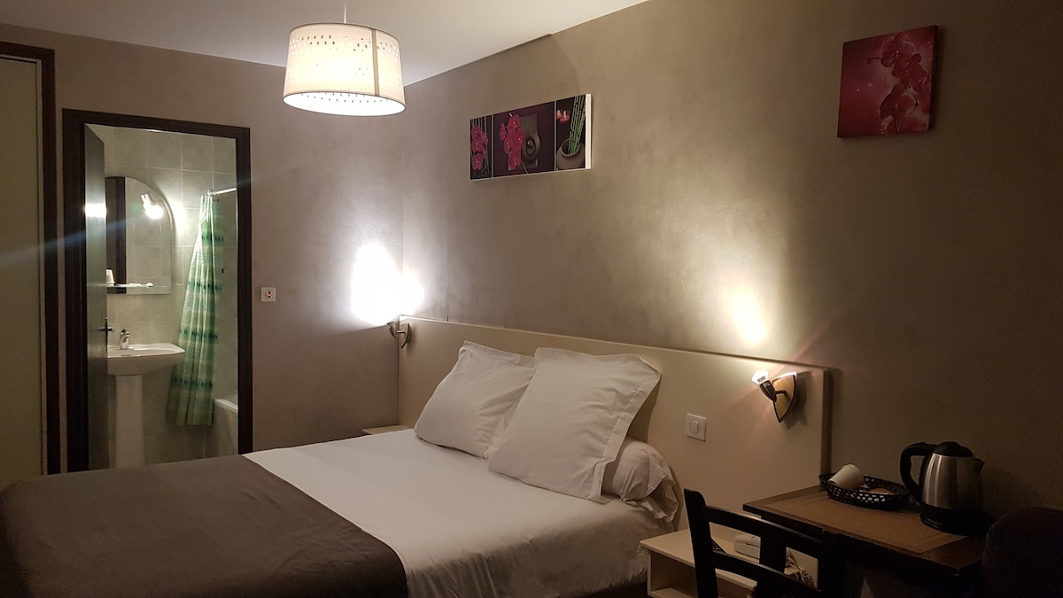 A serene bedroom features a spacious bed with layered white linens and decorative pillows. Soft lighting illuminates the room, highlighting neutral wall tones and floral artwork. A small table with a kettle and cups is visible, while an ensuite bathroom with a glass shower is accessible in the background.