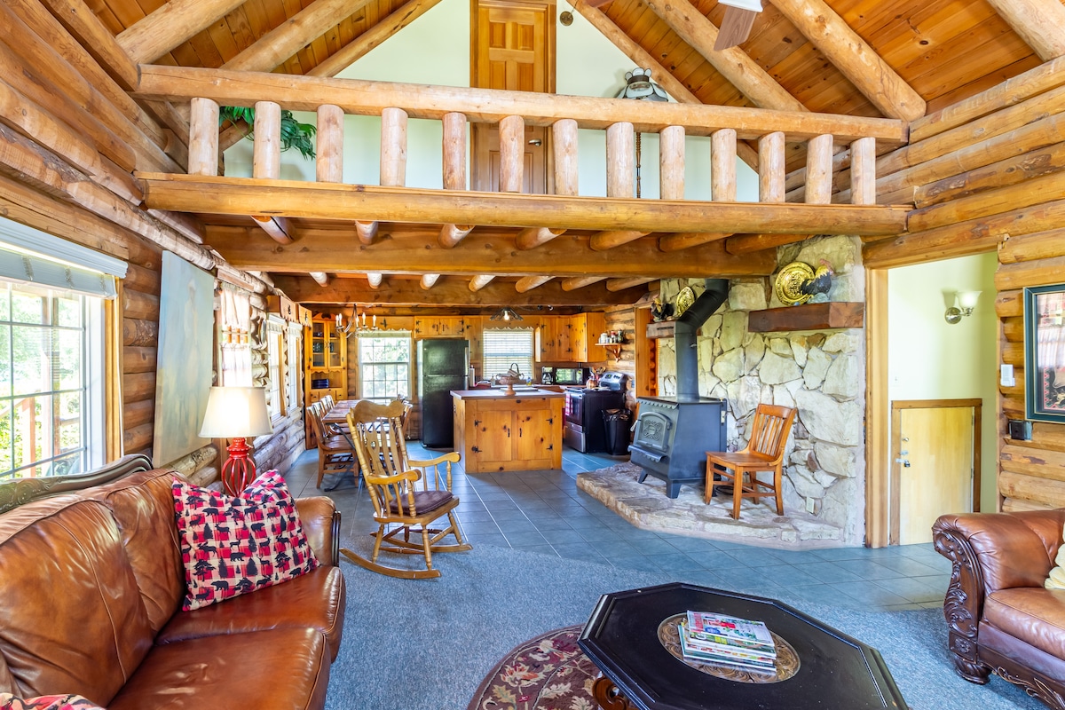 A log cabin's inviting interior features high ceilings supported by hand-hewn beams. A stone fireplace is situated beside a kitchen area, while a cozy seating arrangement includes leather sofas. An upper loft area overlooks the main space, enhancing the cabin's rustic charm.