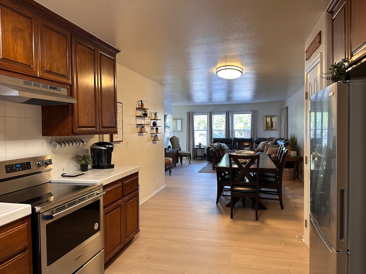 An open kitchen and dining area is seen, featuring warm wooden cabinetry and stainless steel appliances. A dining table with six matching chairs is prominently placed beside a large window, allowing natural light to illuminate the space. The inviting living room is visible in the background.