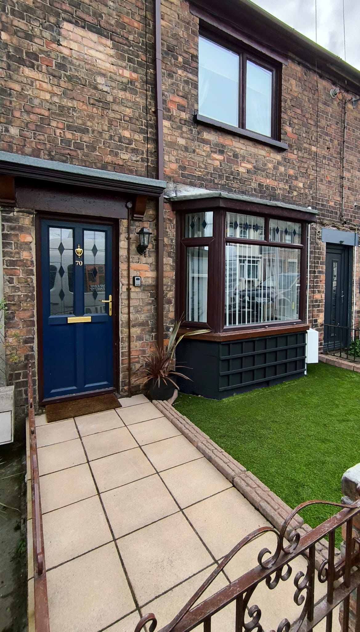 The exterior features a charming entryway with a blue door framed by a decorative porch. A small front garden showcases manicured grass, and the brick facade presents a traditional aesthetic. Windowpanes add character, while the adjacent gate and pathway lead to the entrance.