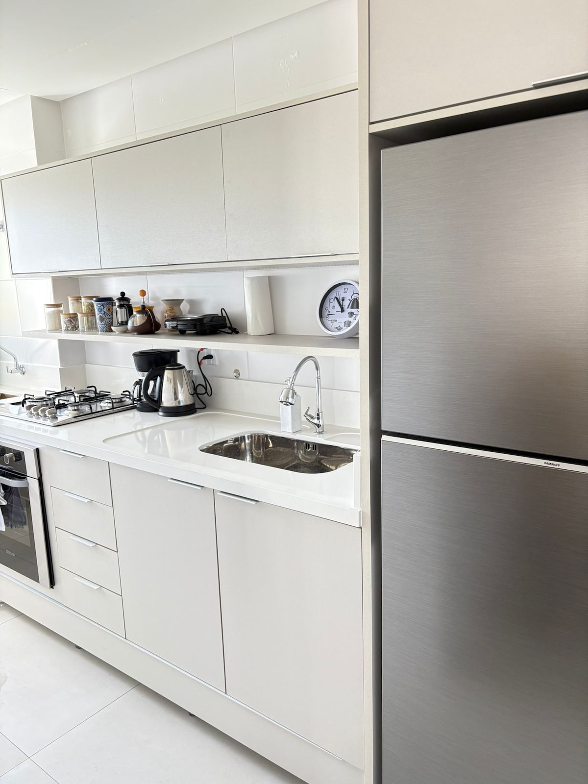 A modern kitchen features sleek white cabinetry and a stainless steel refrigerator. A gas stove and coffee maker are visible, along with a sink and a clock mounted on the wall. Natural light enhances the clean lines and minimalistic design.