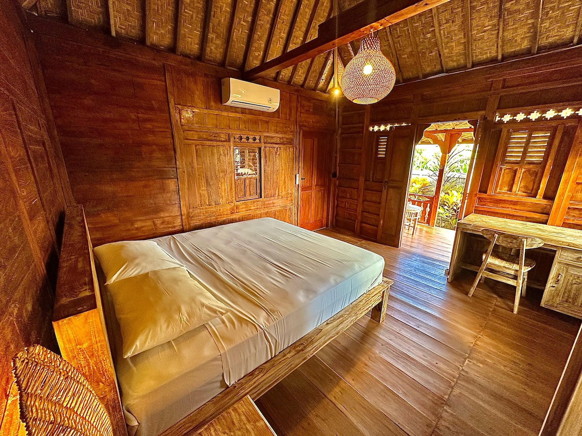 A wooden room features a spacious bed with a light-colored linen spread. Natural light filters through wooden slats, illuminating the rustic decor. A traditional woven chair and a wooden desk complete the room, offering a cozy and functional space for relaxation.