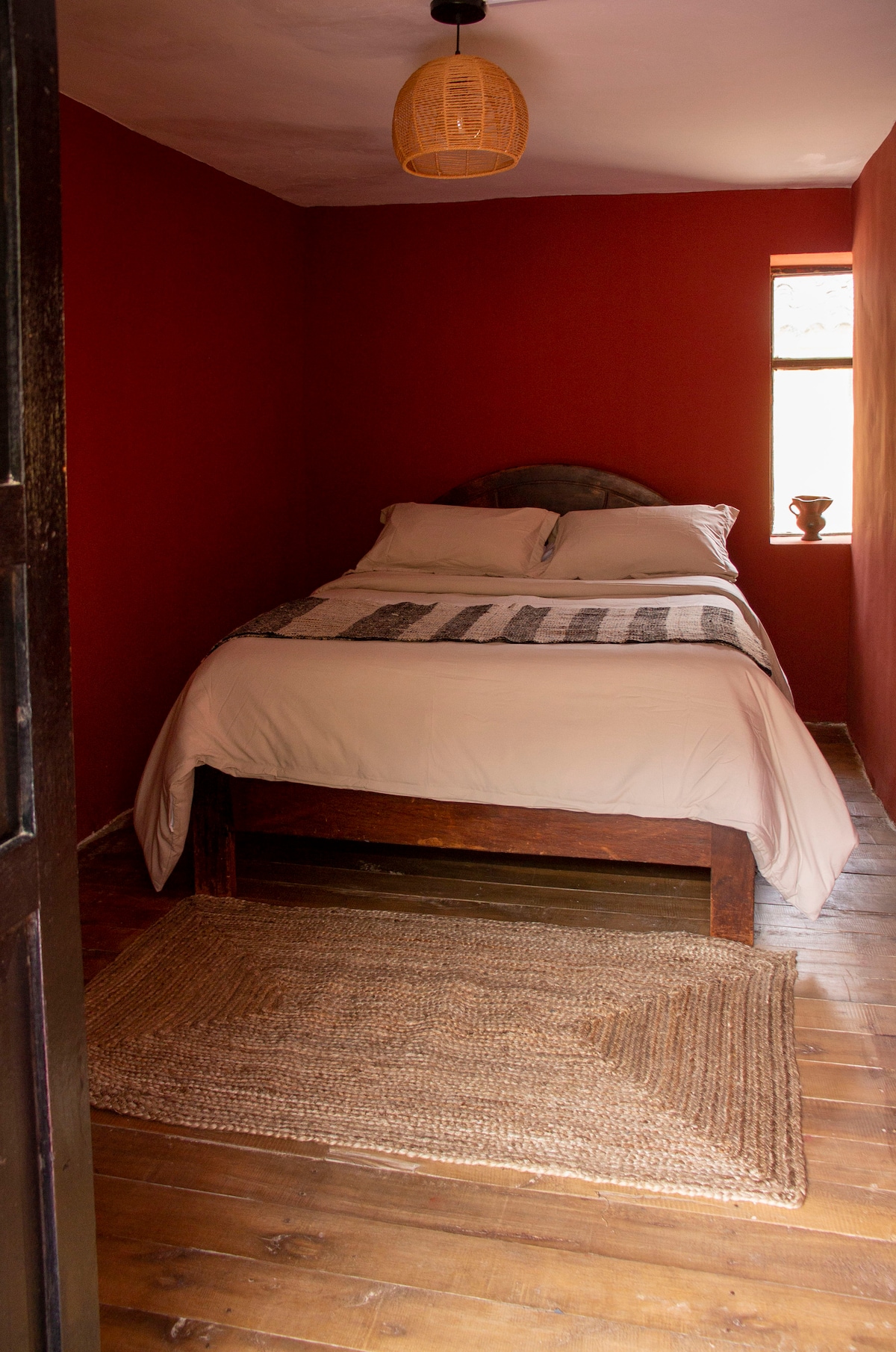 A cozy room features a spacious bed with neutral bedding, complemented by a decorative throw. The warm red walls create an inviting atmosphere. Natural light enters through a small window, highlighting a textured area rug on the wooden floor, adding warmth to the space.