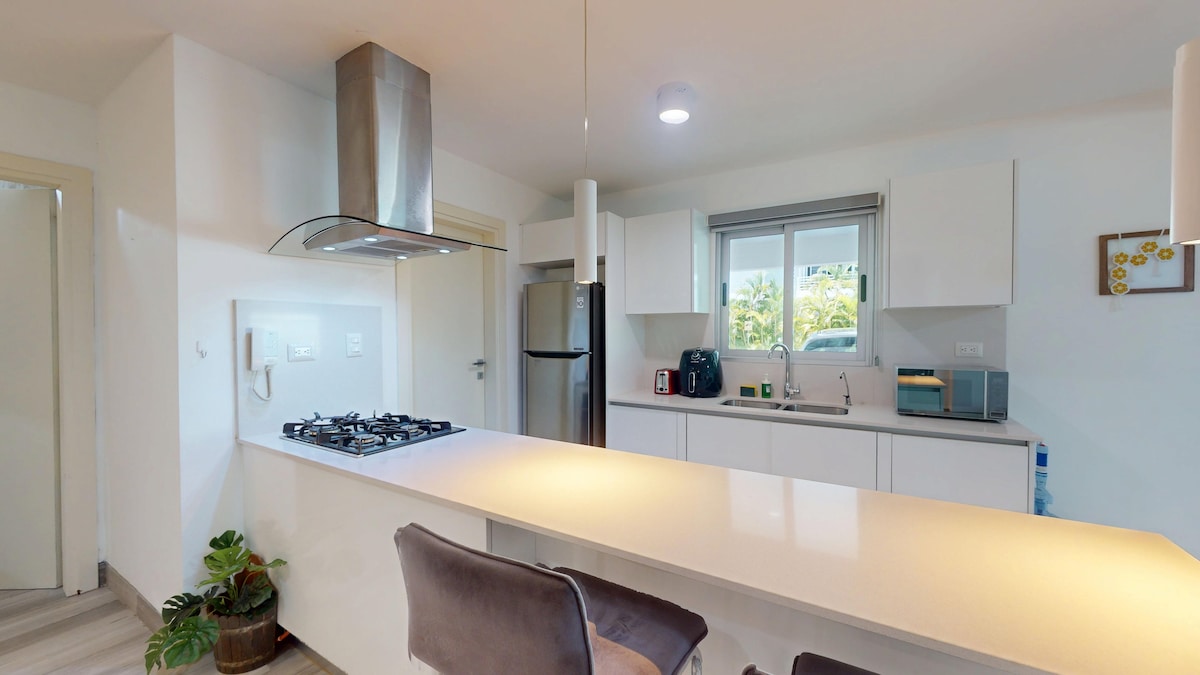 A modern kitchen is presented, featuring sleek white cabinetry and a spacious countertop. A stainless-steel gas stove and hood are positioned prominently. Natural light enters through a window above the sink, framed by views of greenery. An inviting seating area is provided with a dark upholstered chair.