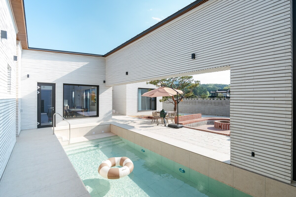 An outdoor area is presented, featuring a comfortable swimming pool with a floating ring. Sunlight filters through the nearby open space, highlighting a patio umbrella and a cozy seating area for gatherings. Surrounding greenery adds a touch of nature to the tranquil setting.