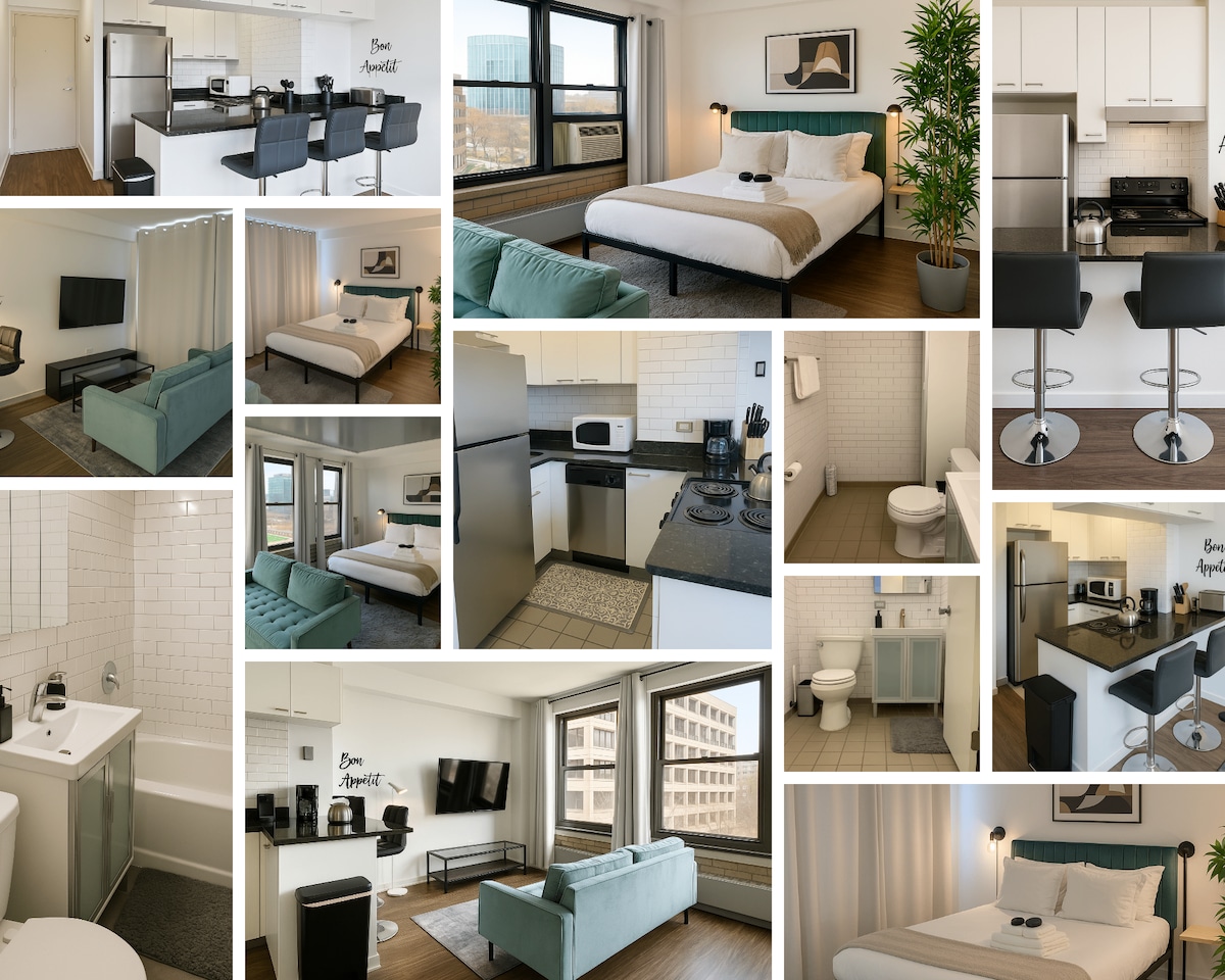 A bright studio space features a full bed centered against a neutral wall. A comfortable sofa sits in front of a mounted TV, while a kitchen area showcases modern appliances and a dining counter with stools. Natural light floods through large windows with sheer curtains.