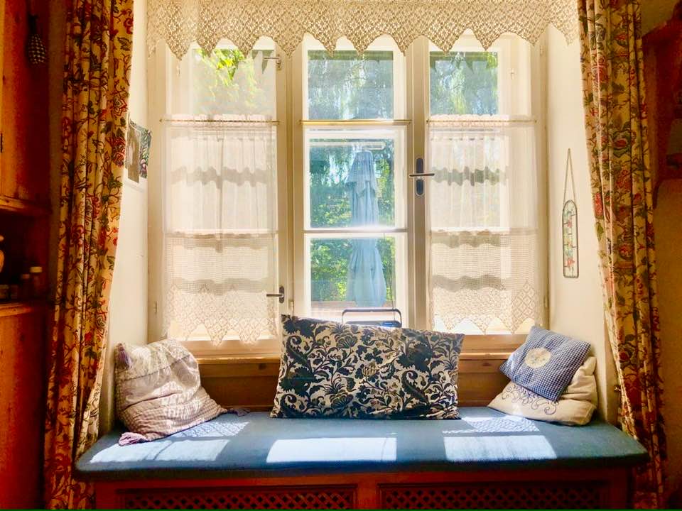 A cozy window seat is framed by light, airy lace curtains, allowing natural sunlight to filter in. Pillows in various patterns are arranged on a dark cushion, complementing the wooden structure beneath. Outside, greenery can be glimpsed through the window, enhancing the serene environment.