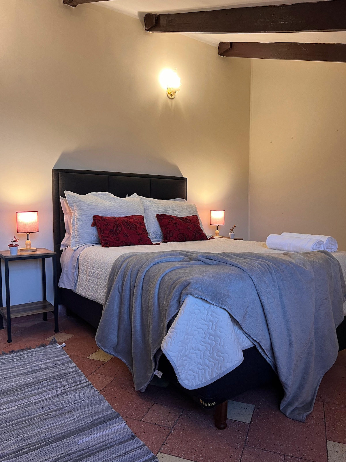 A cozy sleeping area features a spacious bed with a light-colored quilt and two accent pillows. Soft lighting is provided by two bedside lamps. A gray blanket is draped over the foot of the bed, and a textured rug lies on the floor.