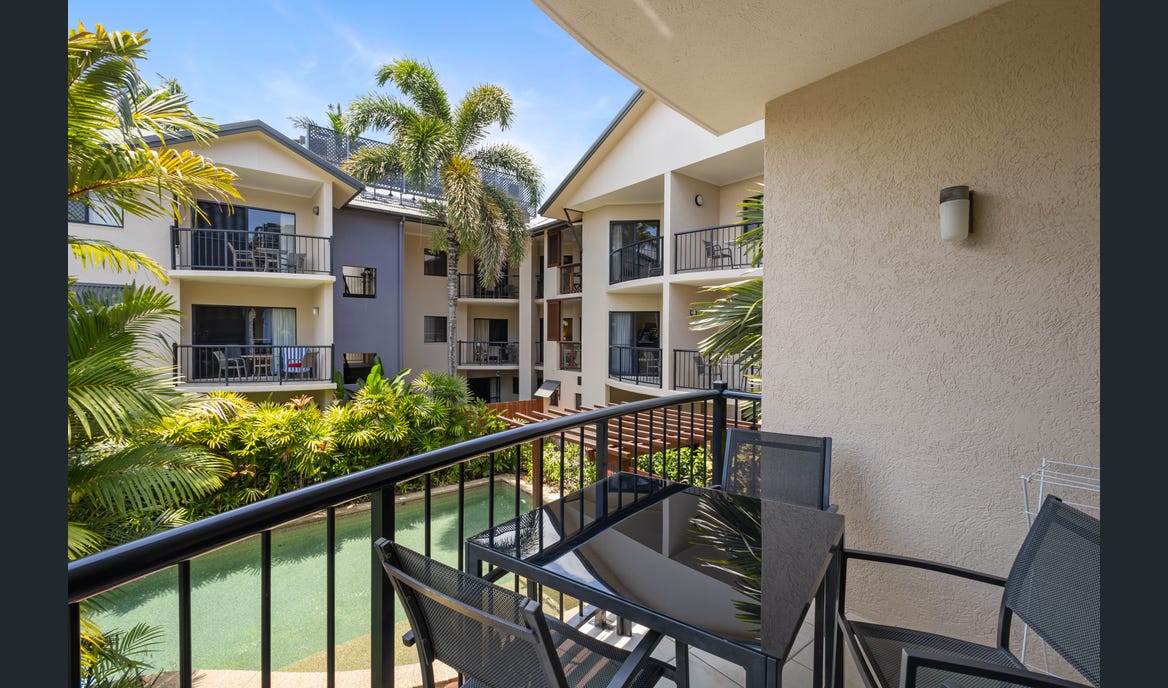 An outdoor balcony features a glass-top table surrounded by four black chairs. Lush tropical plants and palm trees create a serene backdrop, with a glimpse of a nearby swimming pool visible below. The balcony is situated within a multi-level resort structure.