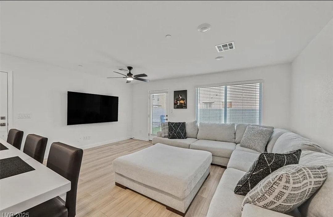 A spacious living room features a soft sectional couch facing a large wall-mounted 75” smart TV. Natural light enters through multiple windows, and a ceiling fan is positioned above. A dining table with dark chairs is visible, enhancing the functional layout of the area.