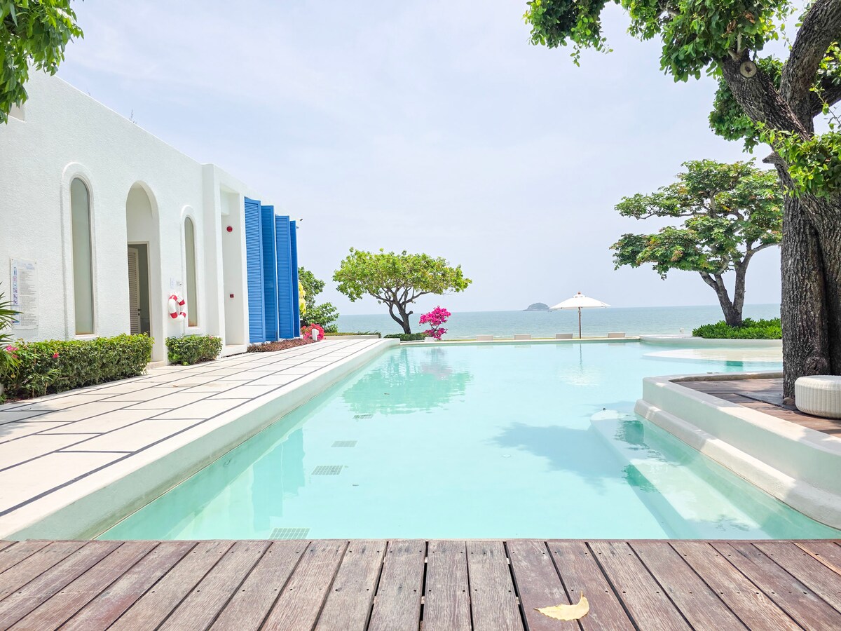 An expansive pool stretches toward a serene beachfront view, surrounded by lush greenery and flowering trees. A wooden deck borders the pool area, enhancing the outdoor space. Blue and white architectural elements create a calm atmosphere, inviting relaxation amidst nature.