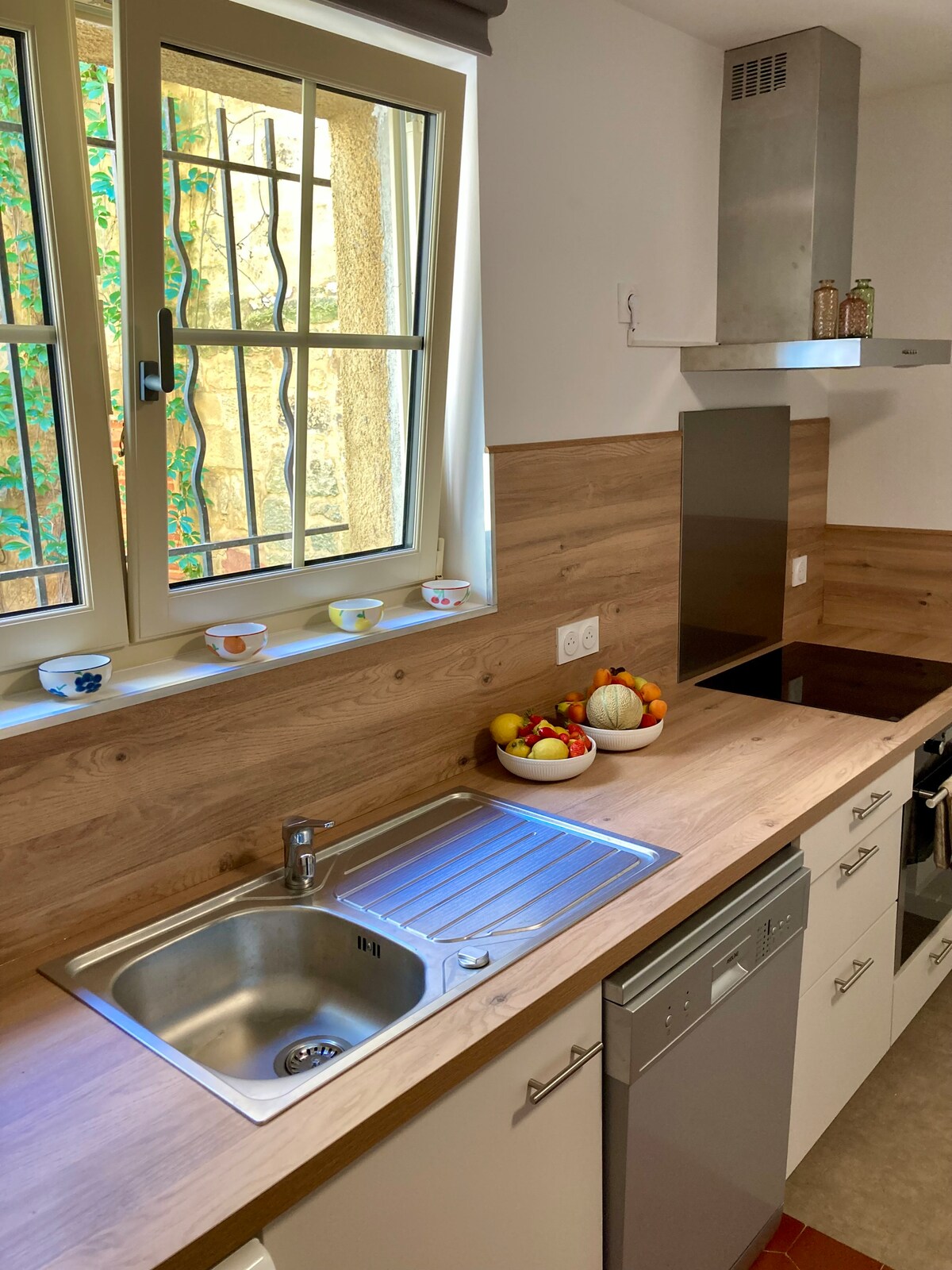 A modern kitchen is presented with wooden paneling and sleek countertops. A stainless steel sink sits beside a dishwasher, with a bowl of colorful fruits displayed nearby. Light filters in through a window with decorative bars, enhancing the inviting workspace.