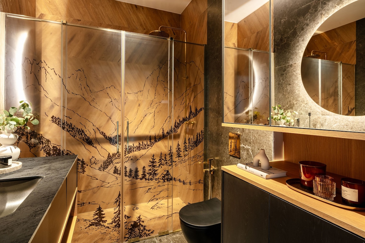 An elegantly designed bathroom features a glass shower enclosure with etched mountain scenery. A sleek dark toilet is visible alongside modern cabinetry that holds decorative items. Warm wood tones and soft lighting create a calming ambiance, complemented by natural elements on the countertop.
