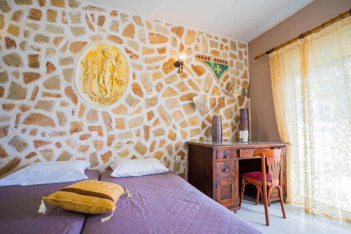 A stone wall design adds character to the room, featuring two spacious beds with white linens. A wooden desk and chair are positioned against the wall, accompanied by decorative elements. Natural light filters through sheer curtains, enhancing the calming atmosphere.