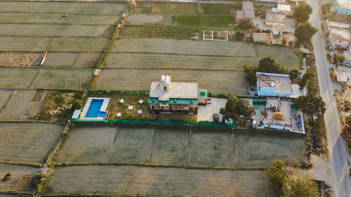 An aerial view captures the resort's layout nestled among expansive fields. A rectangular swimming pool is visible, surrounded by lounge chairs, while the main building occupies a central position, accented by a green roof. Nearby, additional structures and land can be seen.