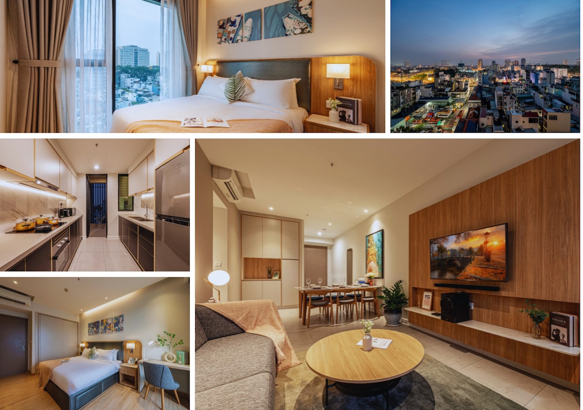 The spacious bedroom features a large bed with soft linens, a bedside lamp, and artwork above it. A large window offers city views. The kitchen area showcases modern appliances and ample counter space. The living area contains a sofa, coffee table, and a TV mounted on a wooden wall unit.