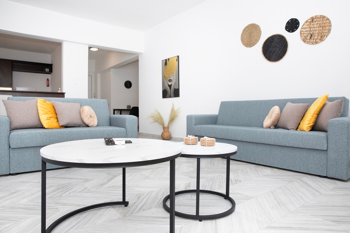 A light-filled living area features two comfortable sofas adorned with decorative cushions. A contemporary coffee table is positioned centrally, complemented by decorative items. Wall decorations made of woven materials add texture to the space, enhancing the overall ambiance.