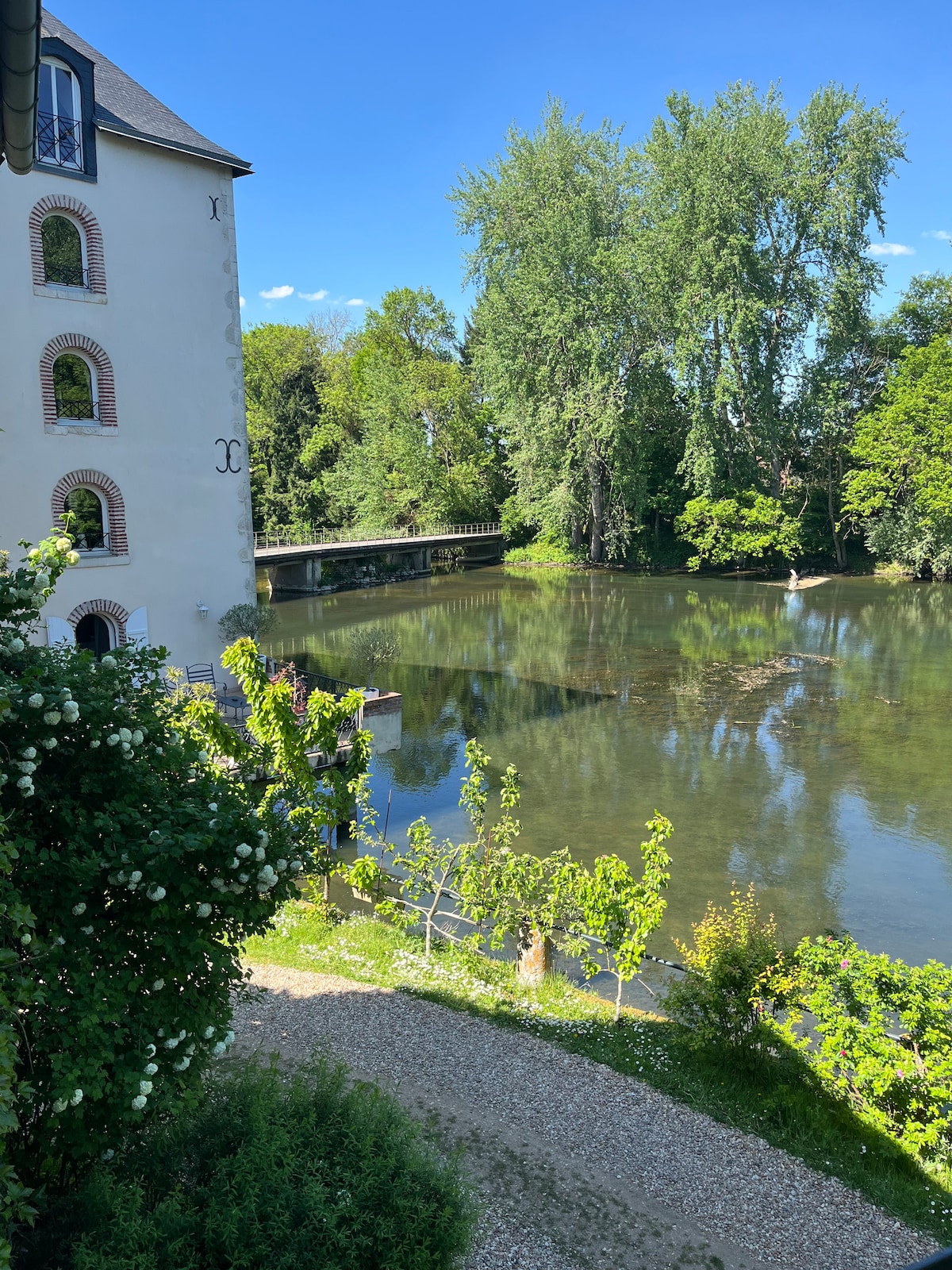 A view of the serene river 'Le Loiret' is displayed, bordered by lush greenery and trees. The calm water reflects the landscape, while a path lined with fresh vegetation leads towards the river. A quaint structure is visible in the background, enhancing the natural setting.