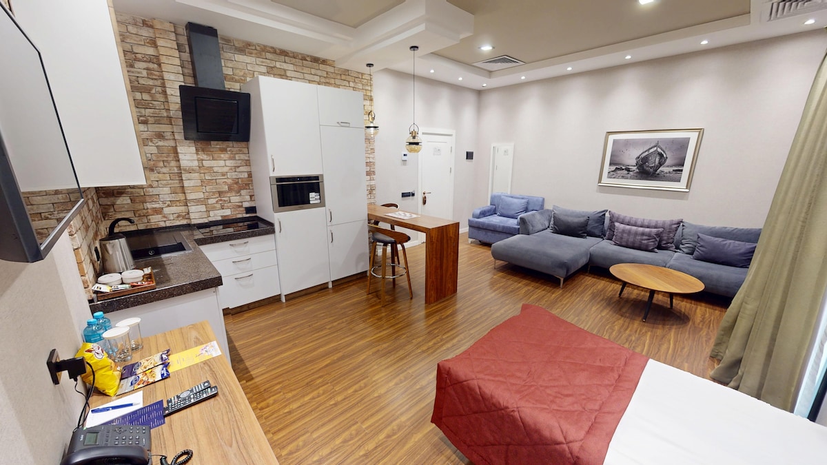 A modern studio space features a spacious bed with a reddish-brown coverlet, a cozy seating area with a large sectional sofa, and a kitchen area equipped with sleek cabinetry. Warm wood flooring complements the neutral walls and accentuates the contemporary decor.