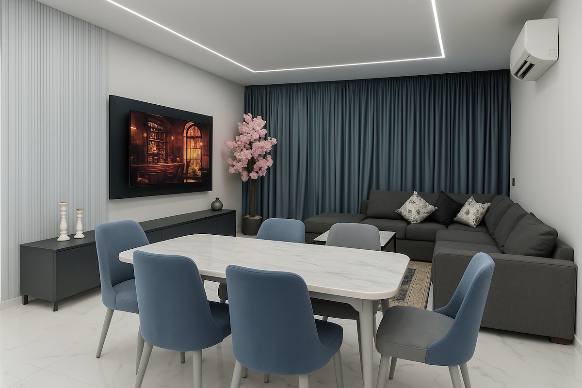 A spacious living area is featured with a dark sectional sofa adorned with decorative cushions. A large marble dining table is surrounded by blue chairs, while a flat-screen TV is mounted on the wall. Natural light is complemented by soft recessed lighting.