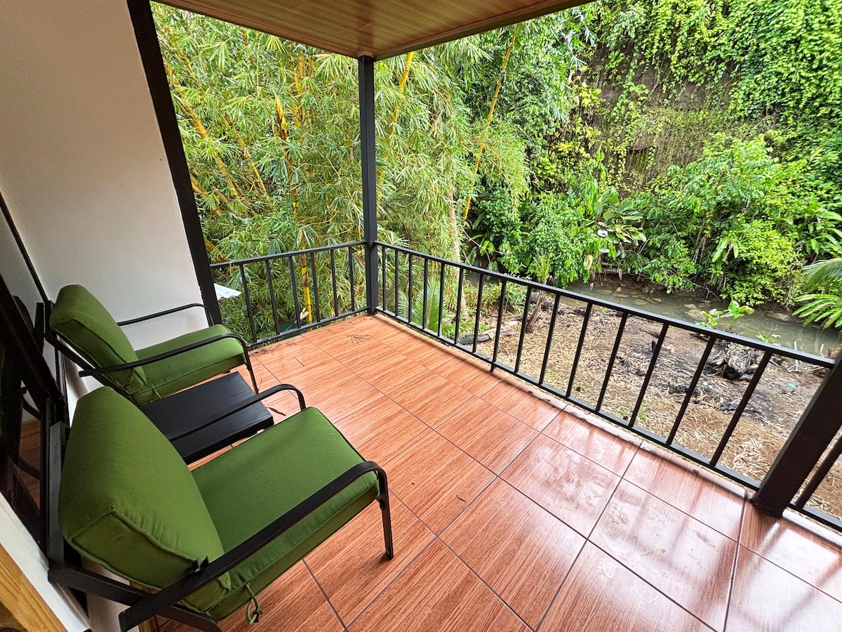 A covered outdoor seating area features two green lounge chairs positioned on a tiled deck. Lush greenery surrounds the space, with a view of a stream and diverse plant life visible beyond the railing.