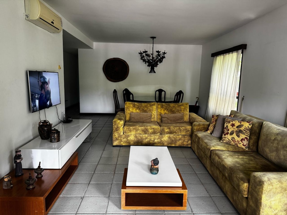 A spacious living area is featured, showcasing two distinct seating arrangements composed of plush sofas in warm tones. A white coffee table sits centrally, while a television is mounted on the wall opposite. Natural light floods the space through large windows adorned with light curtains.