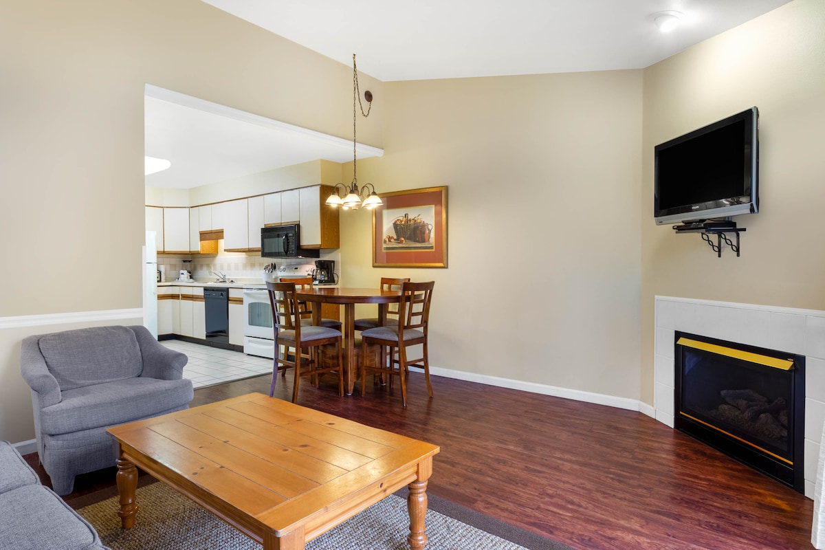 An open living area is displayed, featuring a dining table with four chairs and a cozy sofa. A coffee table sits on a textured rug, while a wall-mounted TV is visible above a modern fireplace. Light-filled kitchen counters and cabinetry can be seen in the background.