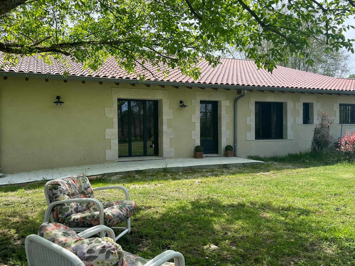 The exterior of the accommodation features a single-story building with a classic tiled roof. There are large glass doors that open onto a grassy area. Two comfortable armchairs are positioned outside, providing a relaxing space, while window shutters frame the dark windows.