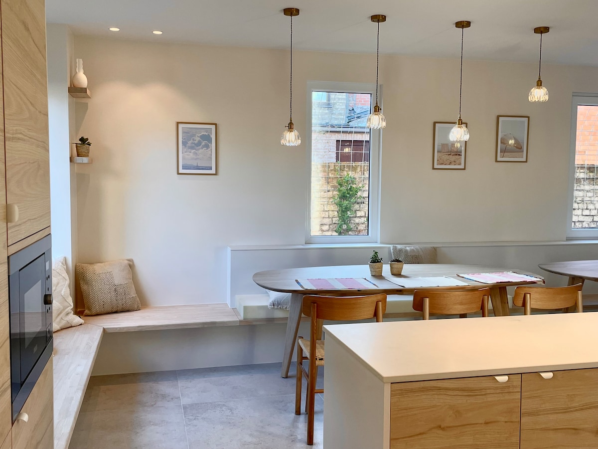 An open kitchen area is featured, with a dining table surrounded by light wooden chairs. A cozy seating nook with a soft cushion is positioned beside a window. Decorative light fixtures hang above, and framed pictures adorn the walls, adding a touch of warmth.