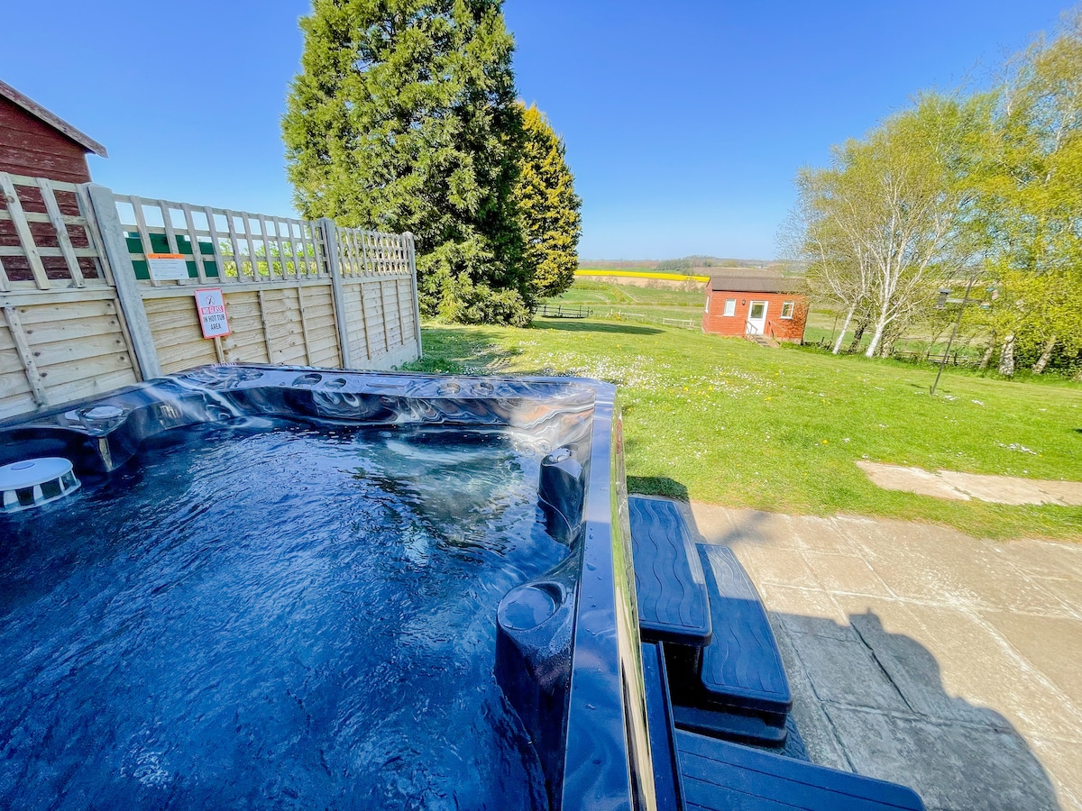A private hot tub sits near a patio area, surrounded by wooden fencing. Inviting views of the expansive green grounds are visible in the background, with a glimpse of a distant building framed by trees and blue skies.