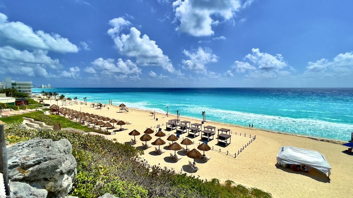 A sandy beach stretches into the distance, showcasing rows of lounge chairs and thatched umbrellas. Gentle waves meet the shore, with bright turquoise waters under a clear blue sky, accented by scattered white clouds.